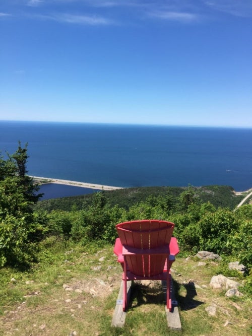 A red adirondack chair sitting in front of a view of forest and ocean.