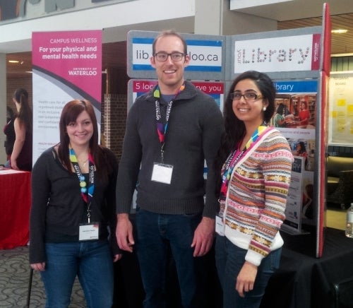 three casual staff members standing in front of the library display board