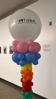 A column of ballons, clusters of ballons forming stripes rising from the ground of green, red, orange, yellow, blue, and pink, with a large white balloon on top branded with the conference logo.