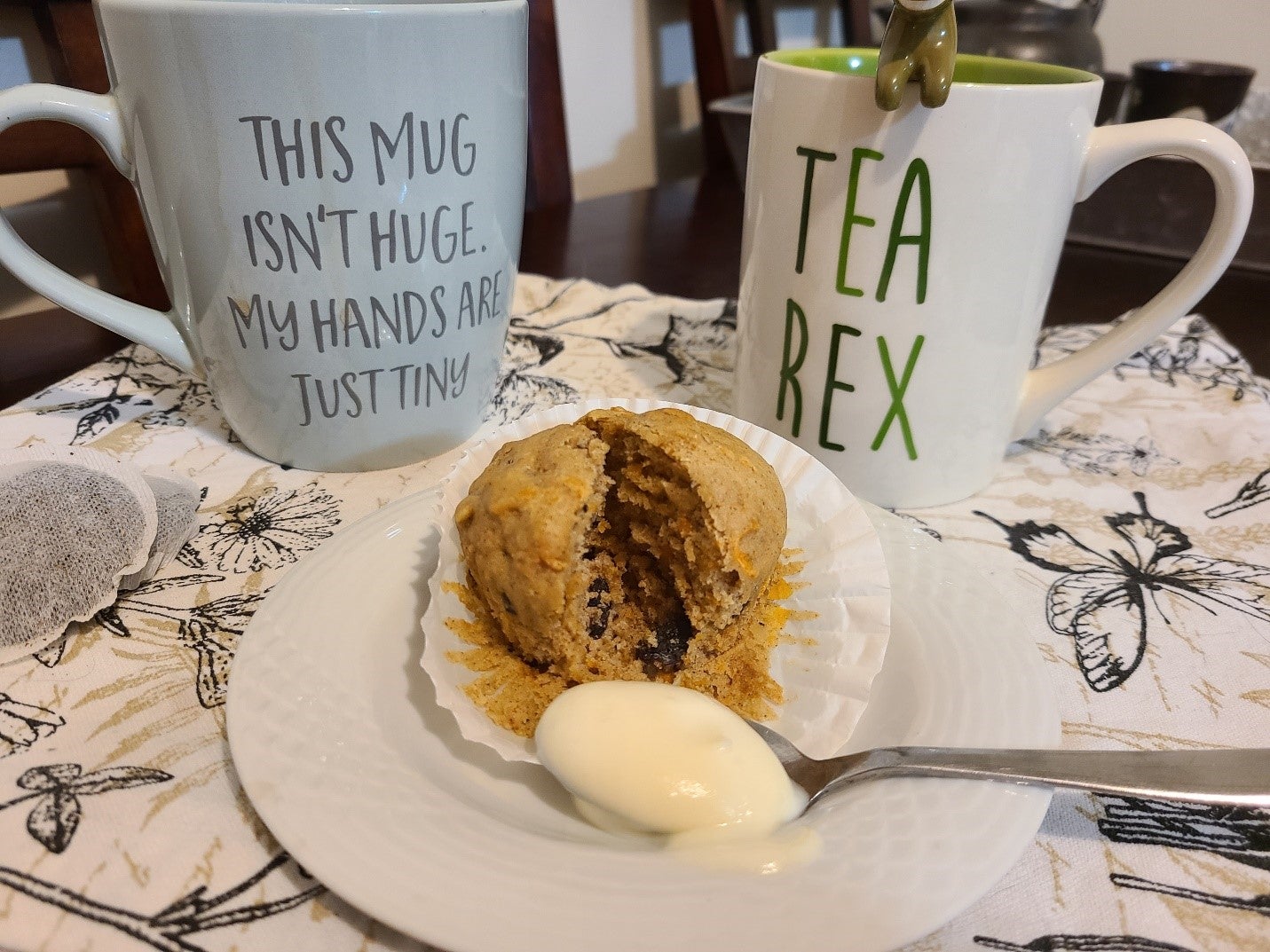 Carrot muffing with a spoonful of vanilla pudding on a small plate. In the background are two mugs of black tea. One reads “This mug isn’t huge. My hands are just tiny” and the other “Tea Rex”.