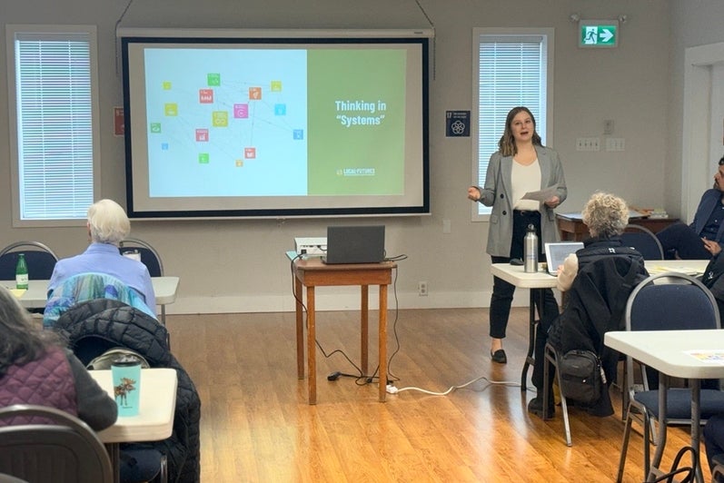 A woman presenting in front of a slide reading "thinking in systems"