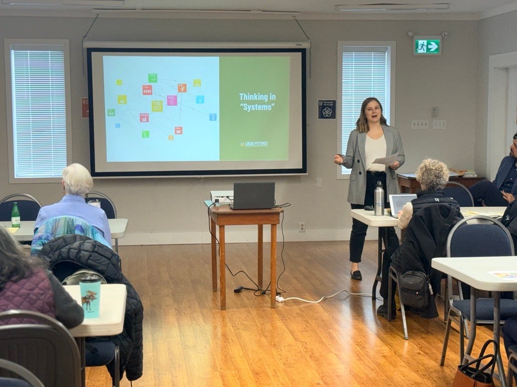 A woman presenting in front of a slide reading "thinking in systems"