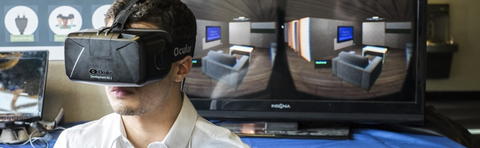 student stands with oculus rift enabled in front of two monitors to show the use of the virtual reality tool.