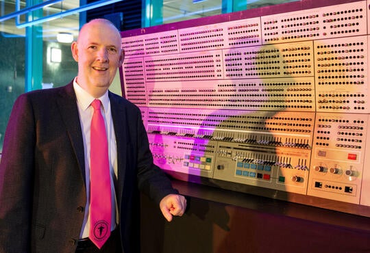man standing beside a large computer wearing a pink tie