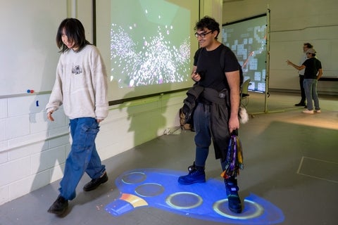 Two individuals standing on and next to blue ocarina projected onto the floor