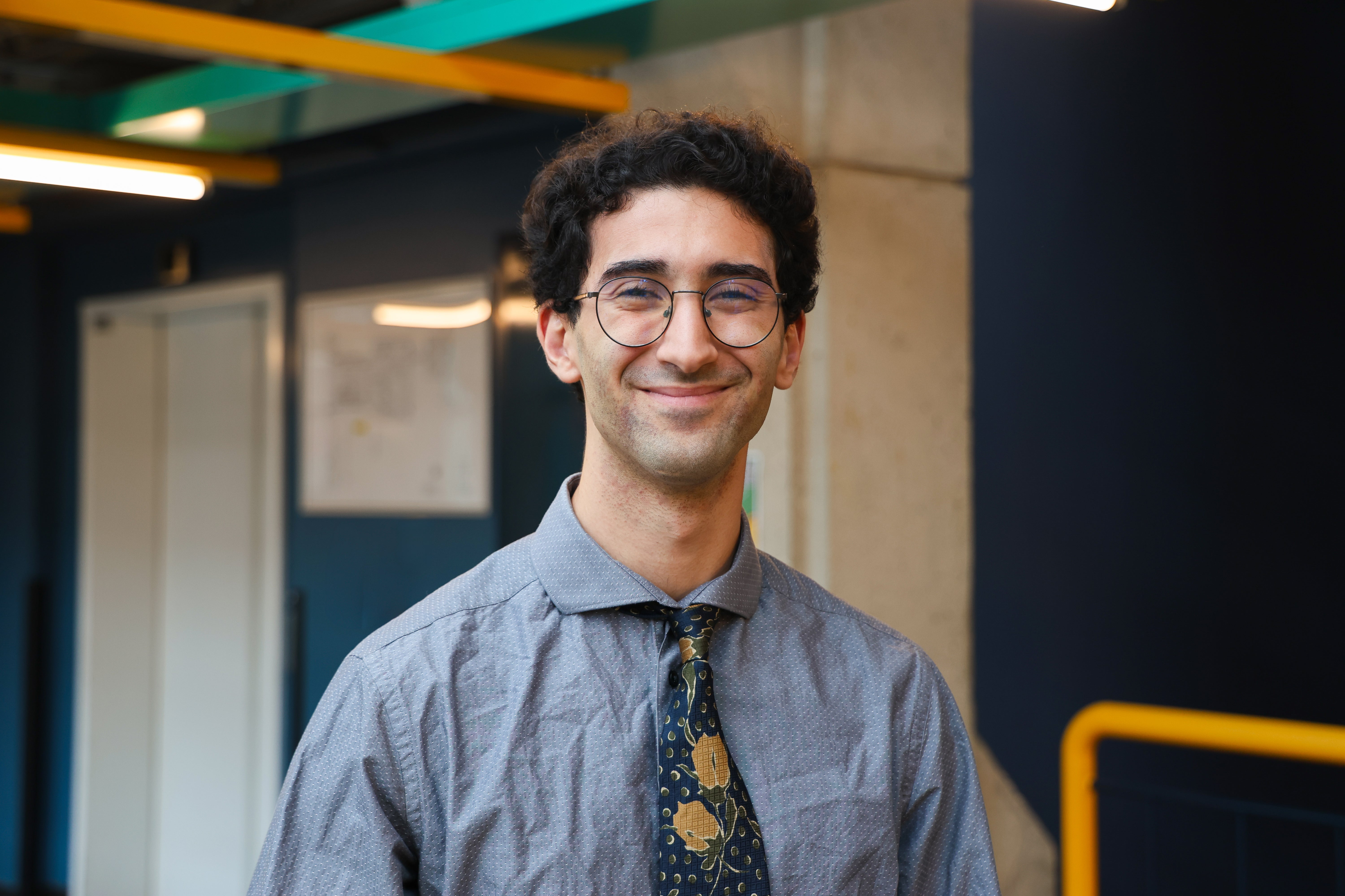 Man with curly brown hair, blue shirt and tie, wearing glasses, facing the camera head on and smiling