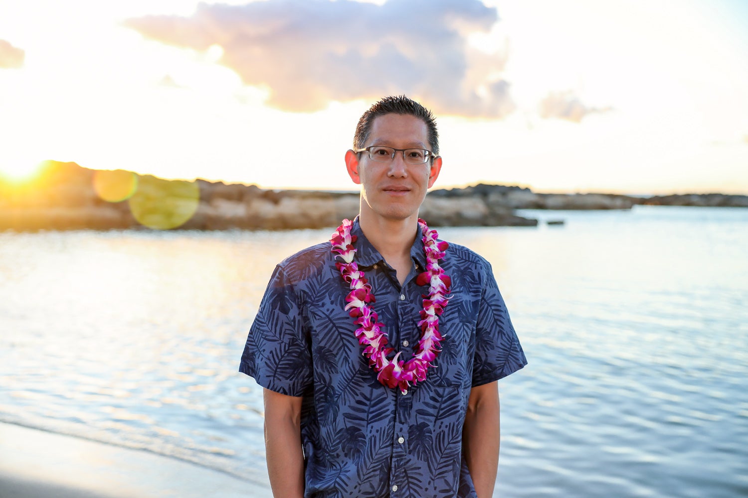 Jimmy Lin, an Asian man with glasses, wears a Hawaiian shirt and lei and stands on a beach