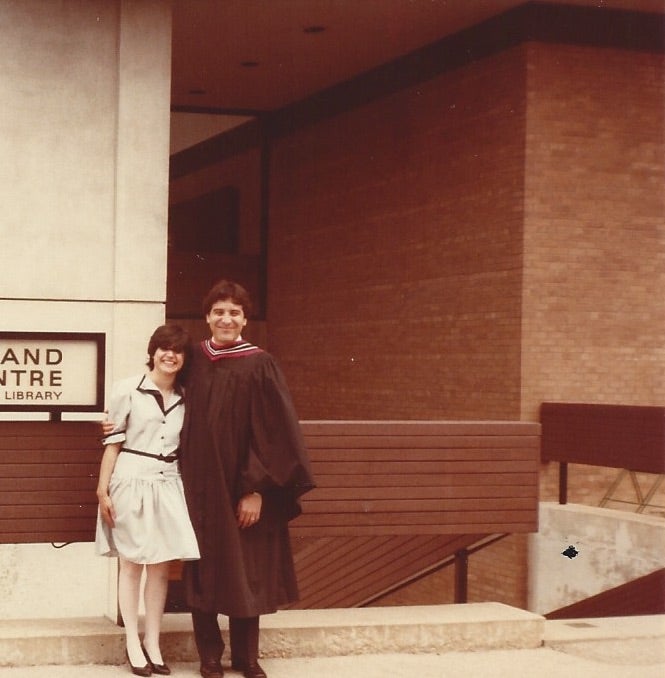 Man in graduation gown stands with woman in front of building.