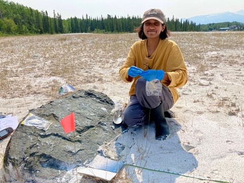 Sana Ahmad collecting rock samples in a field surrounded by trees and mountains. 