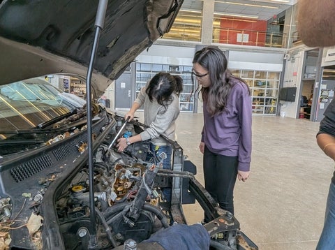 Two students working underneath the hood of a car