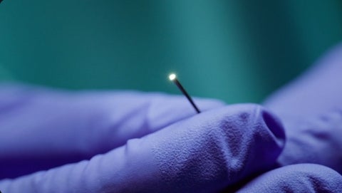 A hand with a blue medical glove holds a Microangioscope, a small camera for looking inside neurovascular, coronary and peripheral vessels.