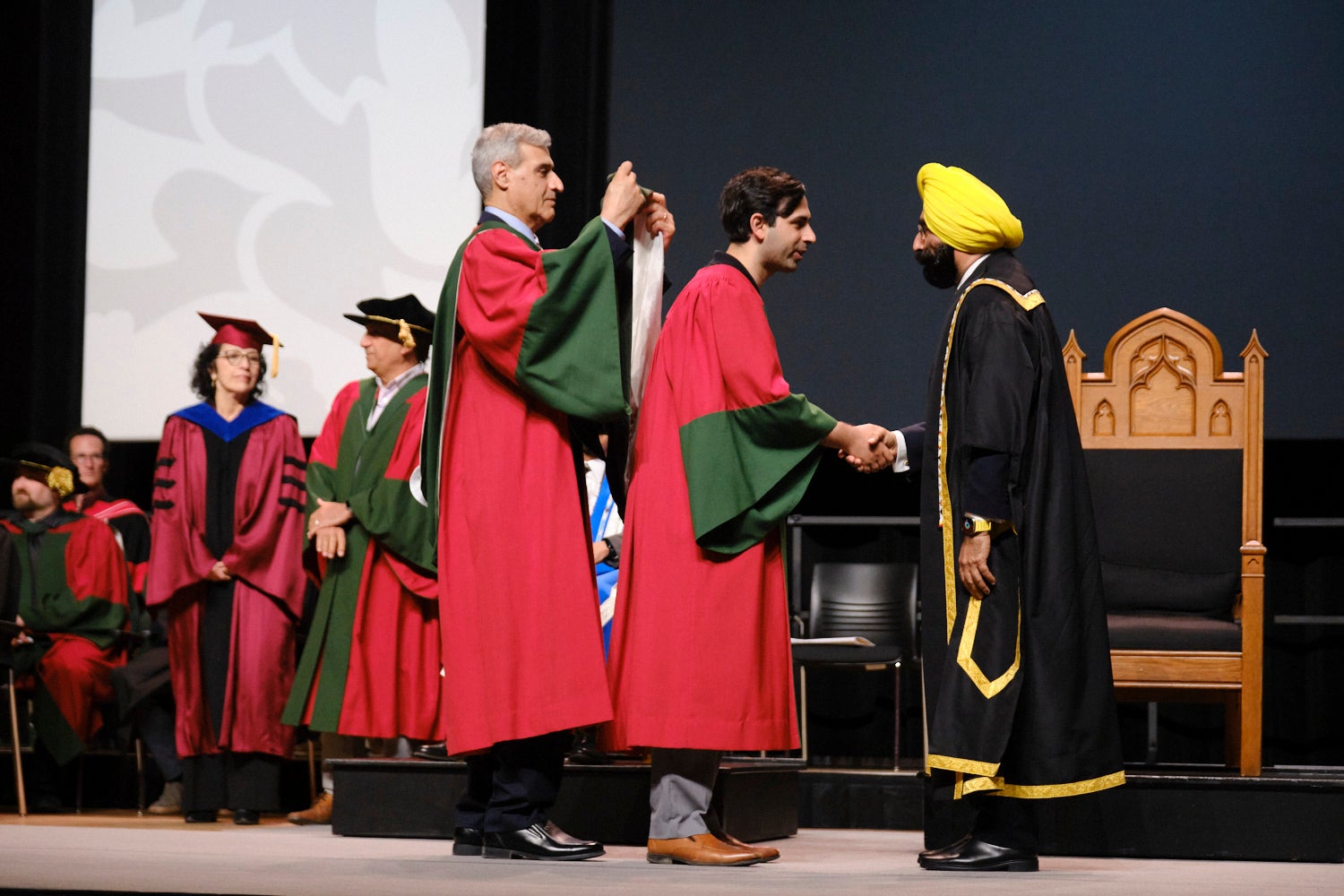 PhD graduate being hooded by Dr. Amir Khajepour, while shaking hands with Waterloo chancellor Jagdeep Singh Bachher. 