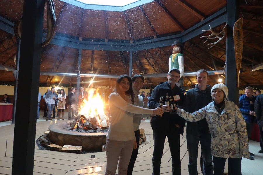 Four people clinking beer bottles in front of a bonfire