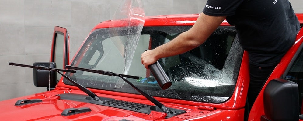 a red car's windshield with a film half on and a arms spraying liquid on the windshield