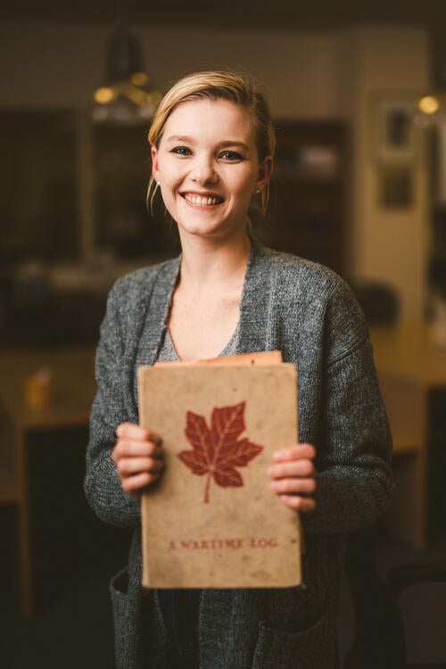 Anna holds a brown notebook with a red maple leaf on its cover