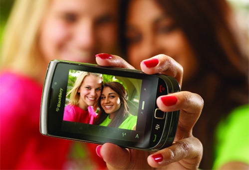 A young woman takes a picture of herself and a friend, who are clearly visible on the screen of a BlackBerry.