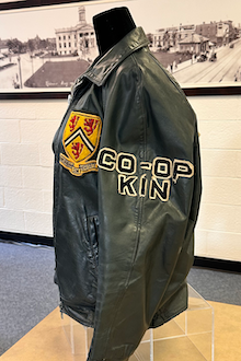 Side view of a black leather jacket with KIN and CO-OP patched on the sleeve and a Waterloo logo patch on the front