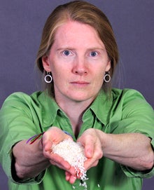 A serious-looking woman in a green shirt holds out her hands, which are full of rice, and through which a few grains of rice are slowly pouring.