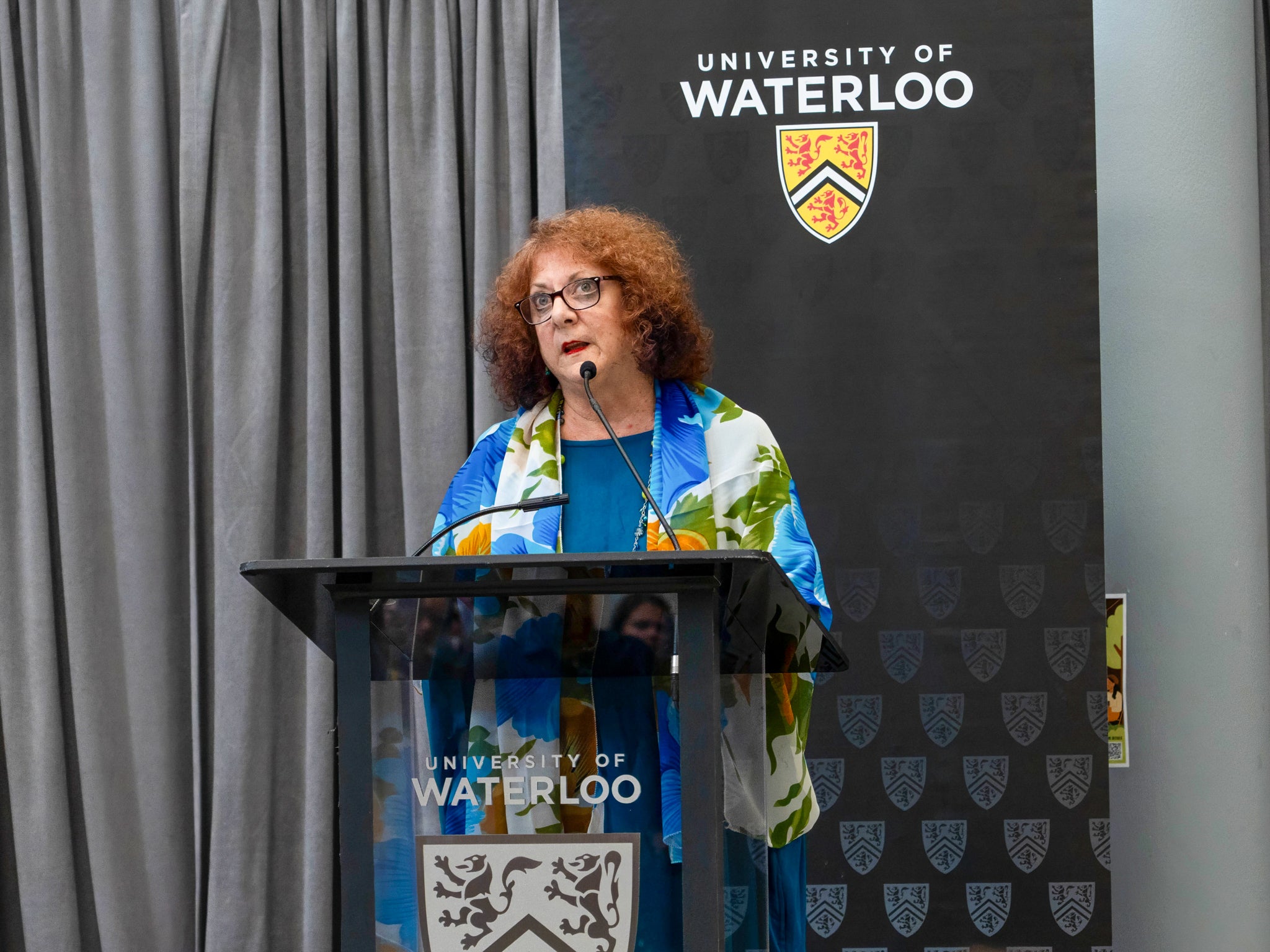 Dr. Sheila Ager speaking at a podium inside Hagey Hall Hub with a University of Waterloo banner behind her