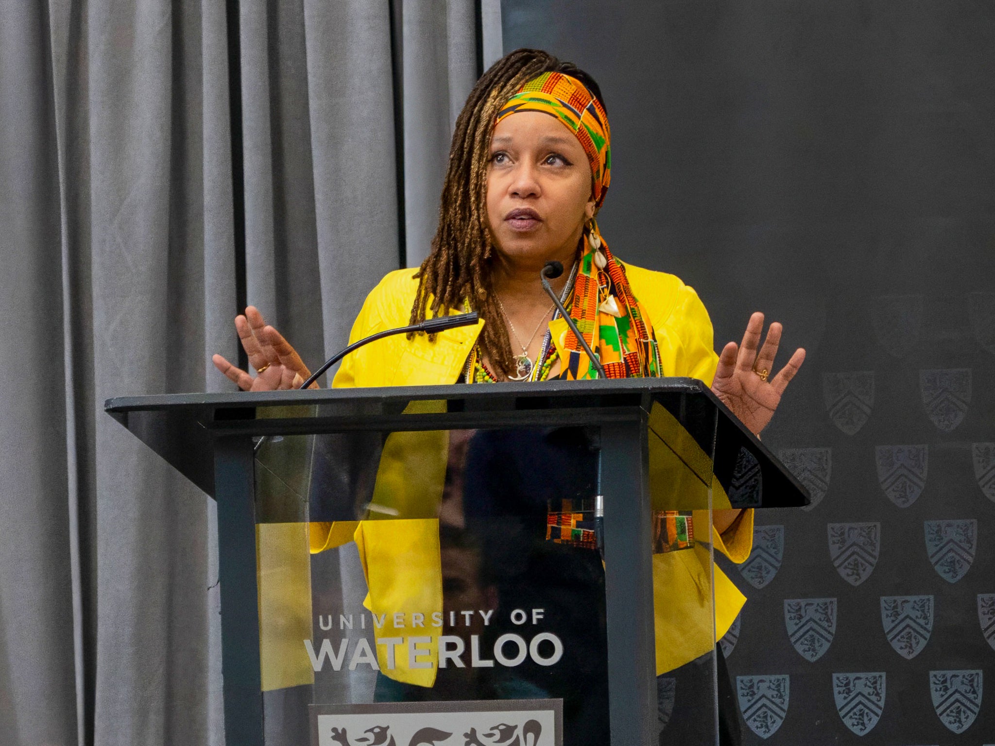 Dr. Laura Mae Lindo speaking at a podium inside Hagey Hall Hub with a University of Waterloo banner behind her