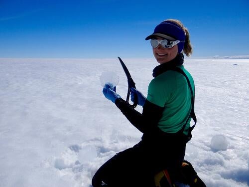 Woman kneels on glacier holding a piece of ice and a saw