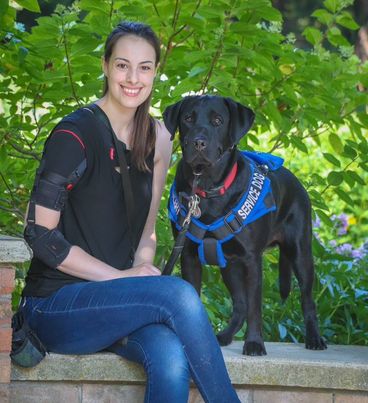 Emma, a woman with brown hair and a shoulder brace, sits next to her service dog. The dog is a black lab wearing a blue harness.