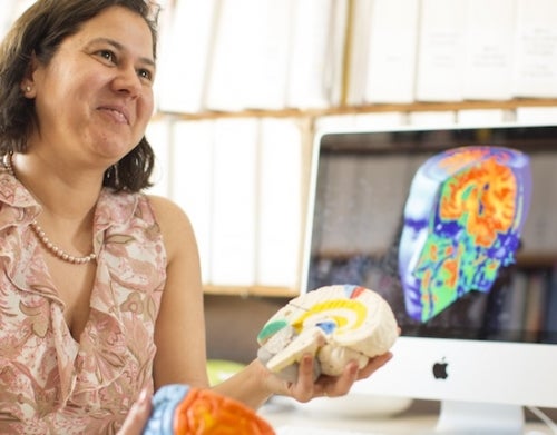 Myra Fernandes, holding coloured models of a brain in front of a computer screen with a different coloured model on it.