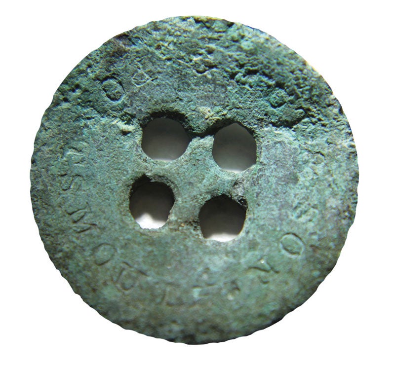 A metal button with letting around the edges. The button is so old tha the metal has turned green an is pitted