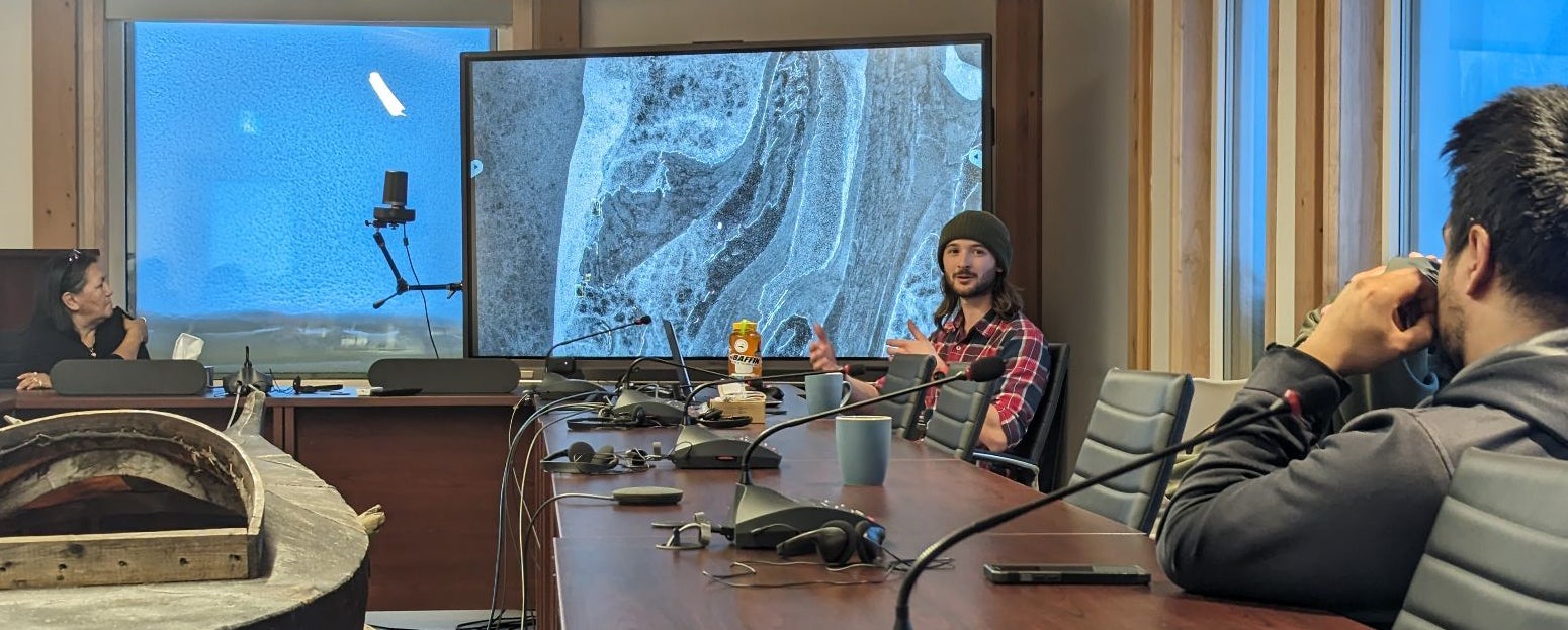 Three people siting at boardroom table with one talking and presenting information on a screen