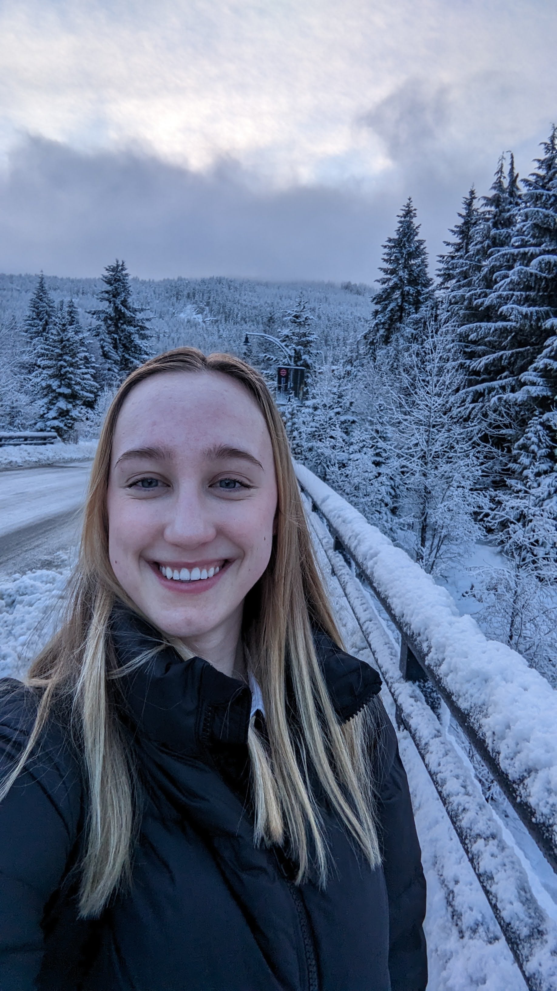 Sarah Hardy has blonde hair and blue eyes. She is wearing a black winter coat and is standing on a snow covered bridge.