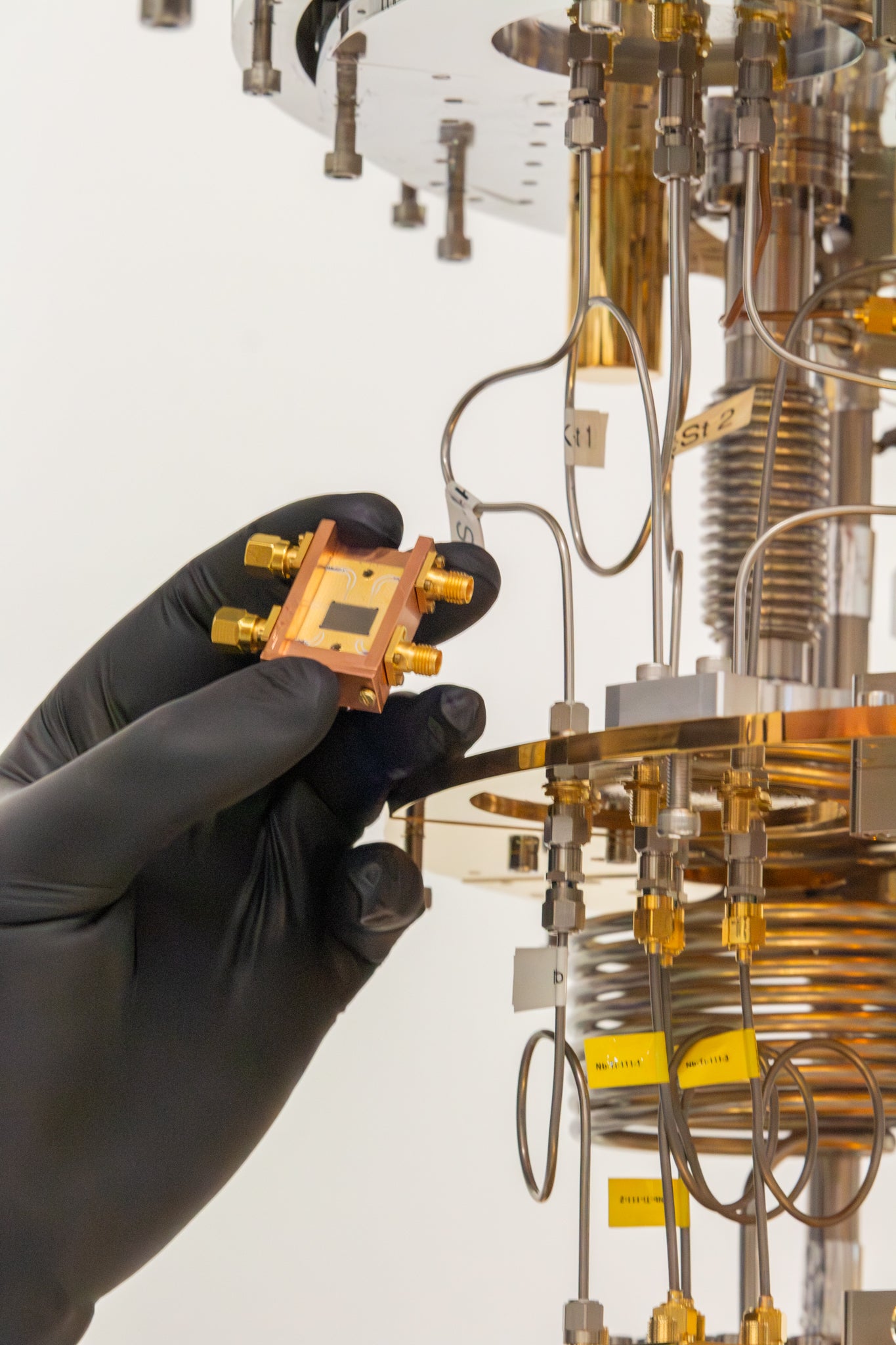 A gloved hand holds a small gold-plated square microchip with four terminals, in front of a laboratory setup with copper coils and tubing.