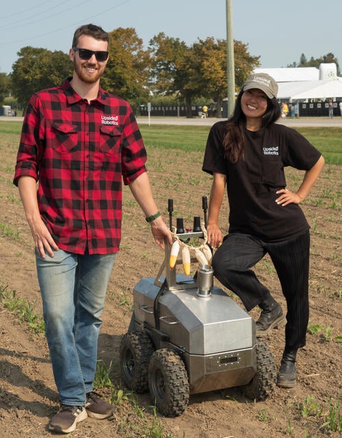 Two people stand in a farmer's field beside a small four-wheeled robot