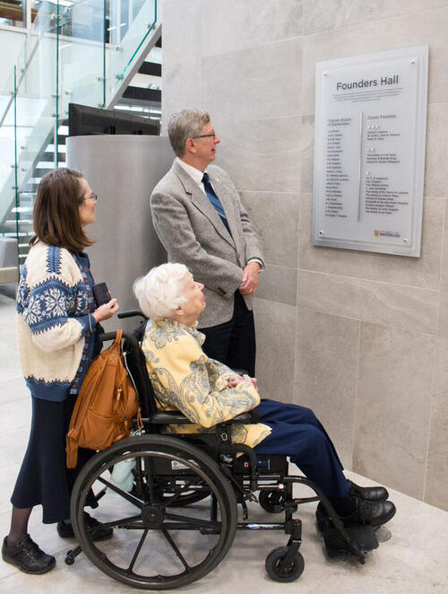 A woman, a man and a woman in a wheelchair look at a wall plaque