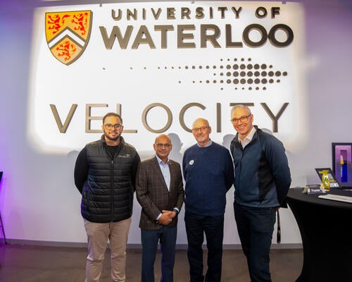 Moazam Khan, Vivek Goel, Mike Stork and John Dick pose in front of the Velocity and UWaterloo sign inside the Innovation Arena