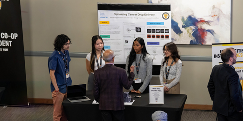 Four students stand at a booth, in front of their i-Capstone poster, talking to a person at a University of Waterloo event.