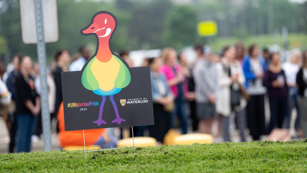 Pride signage featuring a rainbow goose is pictured on a lawn in front of a large group assembled in lot H