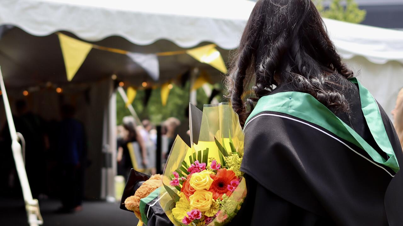 back of graduate wearing robes, green hood and carrying flowers