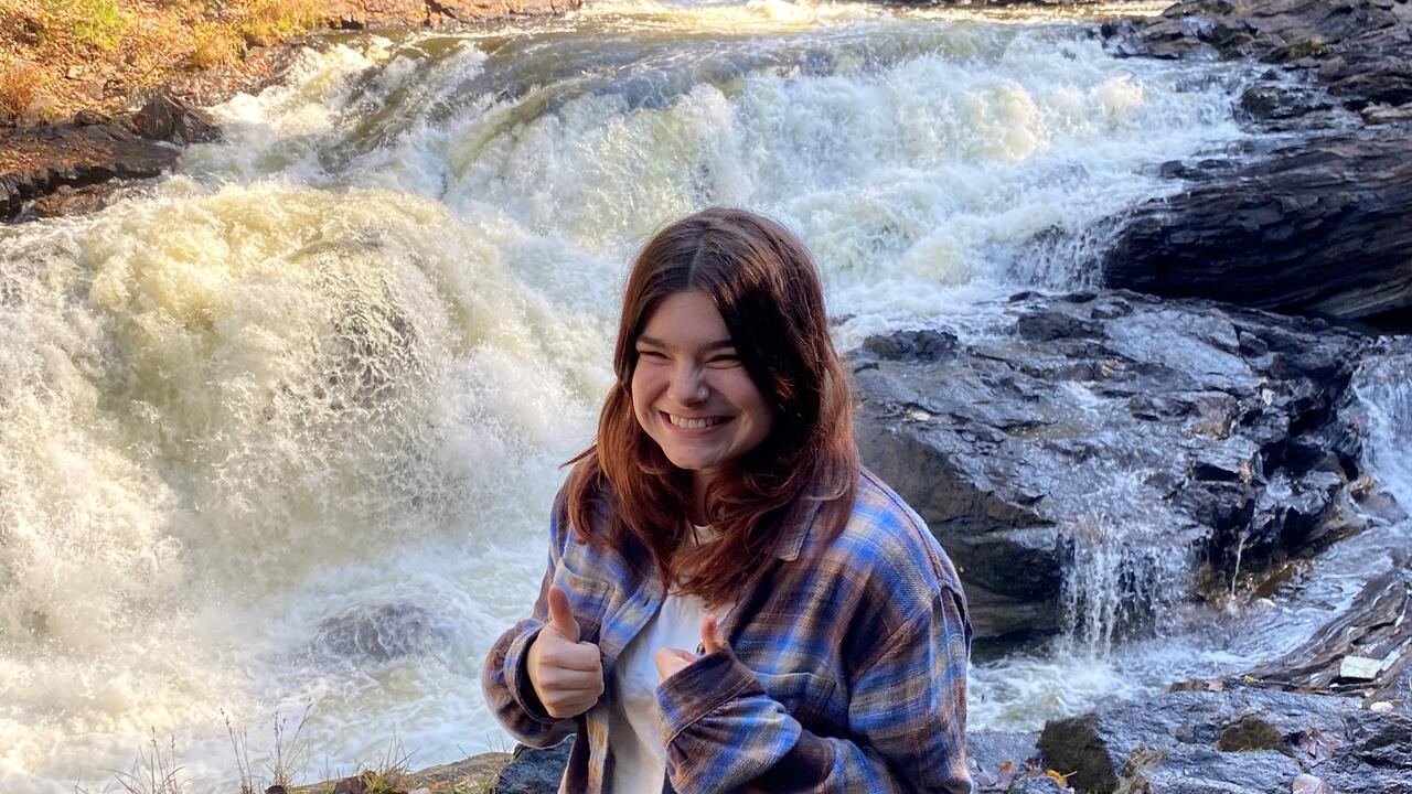 A smiling young woman wearing a  plaid shirt stands in front of a mountain stream