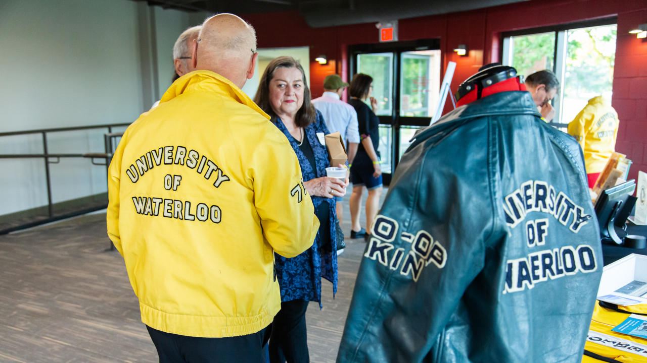 An alum wearing a vintage University of Waterloo jacket, standing beside one on a mannekin