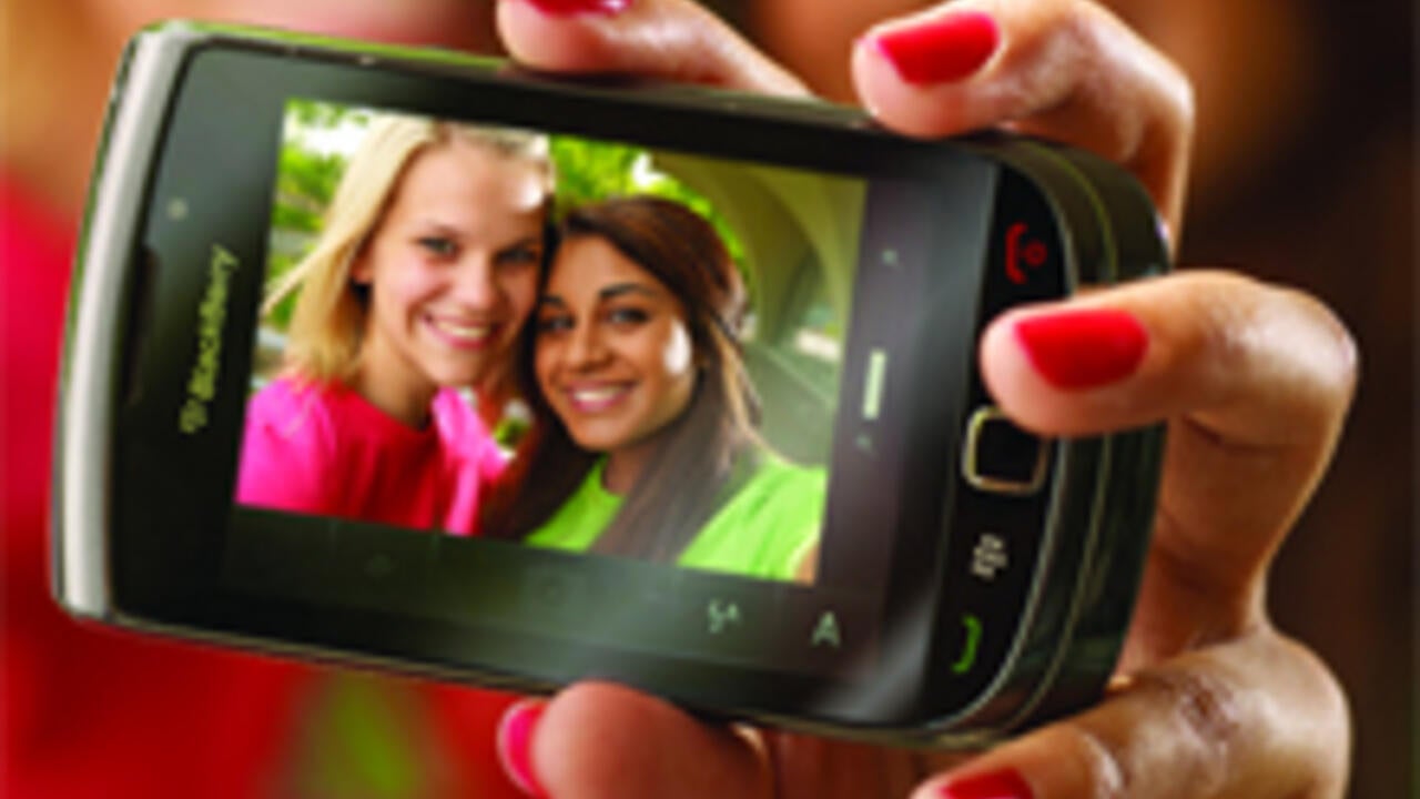 A young woman takes a picture of herself and a friend, who are clearly visible on the screen of a BlackBerry.