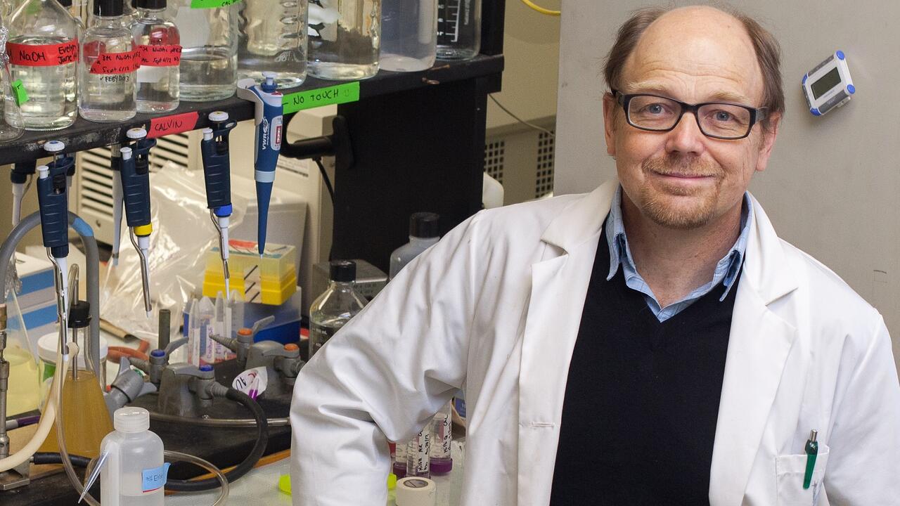 Professor wearing lab coat stands in front of a shelf of beakers in a laboratory
