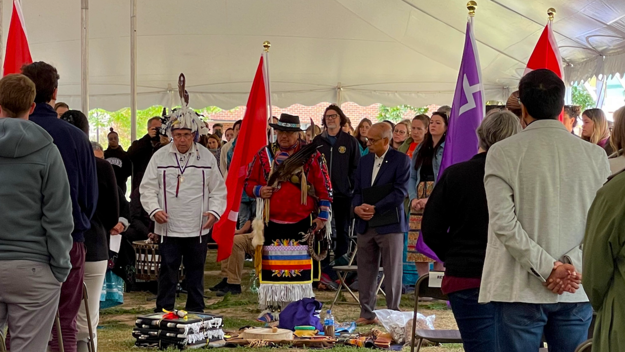 Elder Bill Woodworth, Elder Myeengun Henry and Vivek Goel stand in front of a blanket with traditional Indigenous gifts
