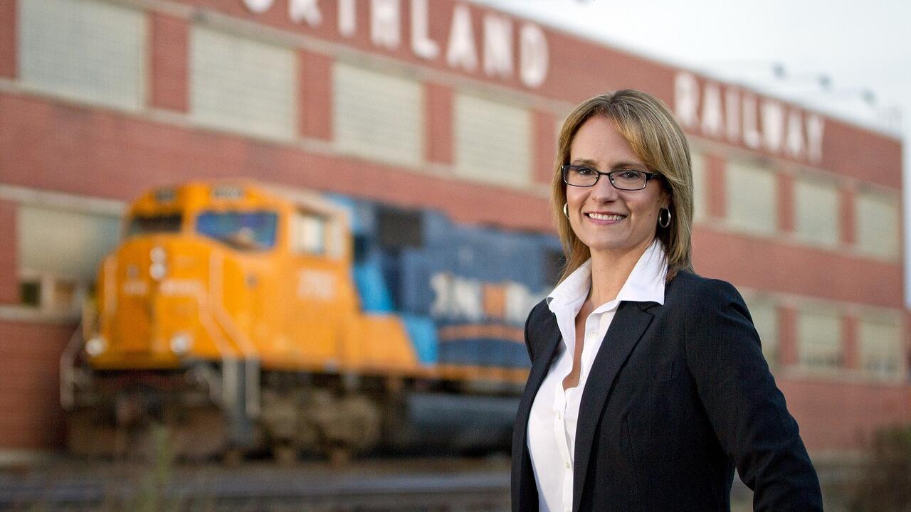Corina Moore stands in front of stopped train on a railway and Northland Railway building