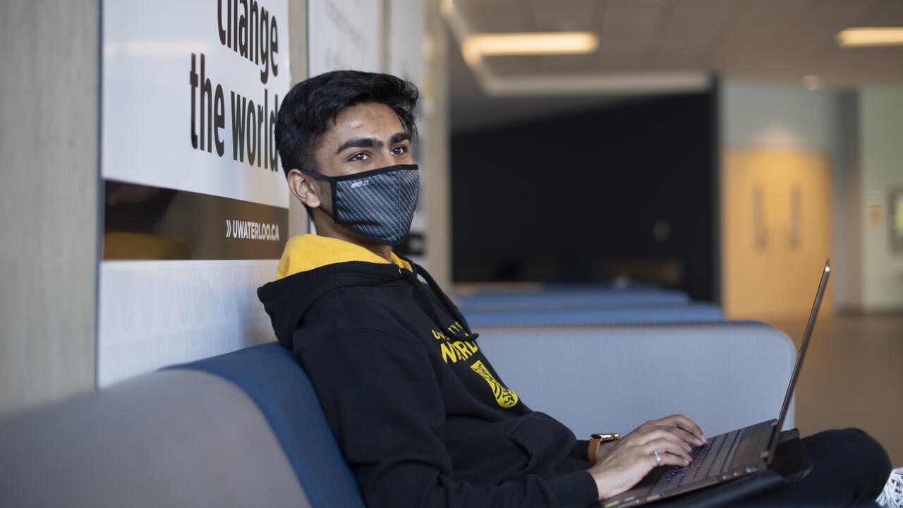 A student sitting in a Waterloo hoodie and mask while typing on a laptop