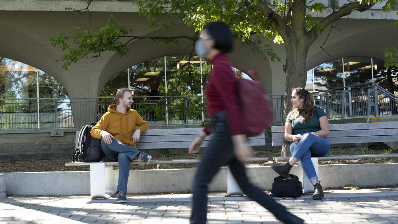 Two students talk while sitting on a bench as another student passes by