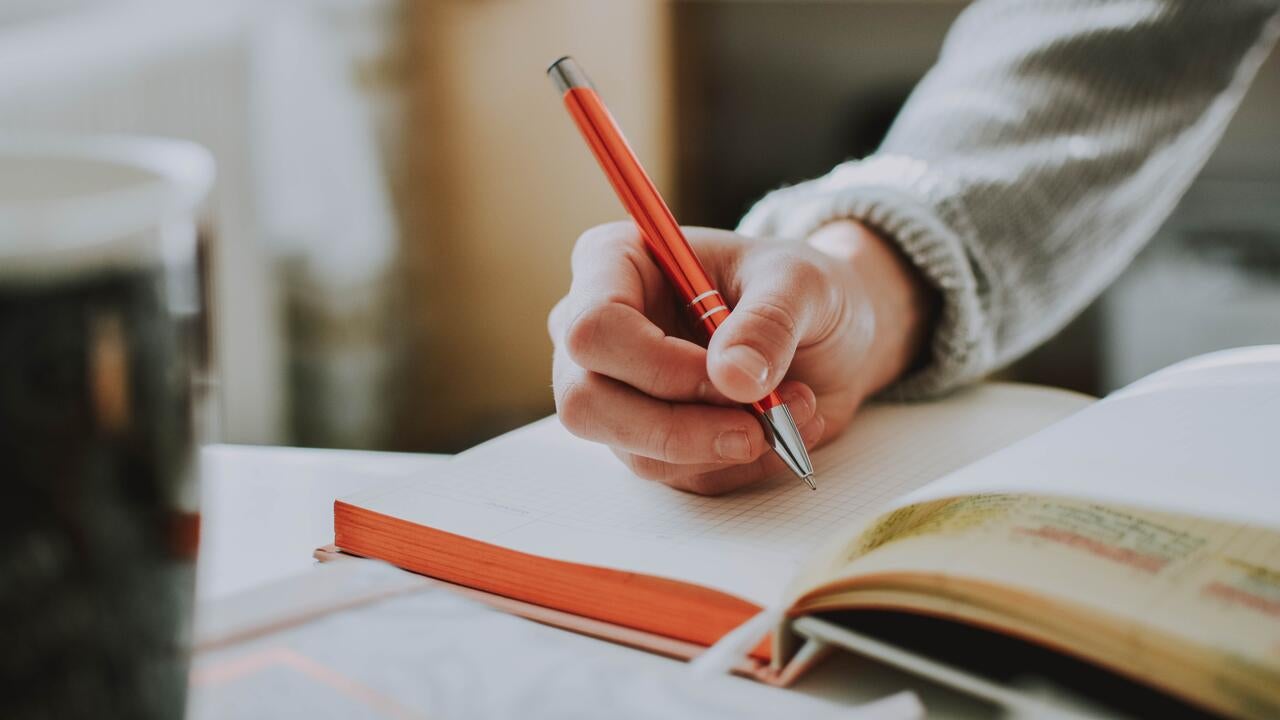 A hand writes in a notebook with an orange pen