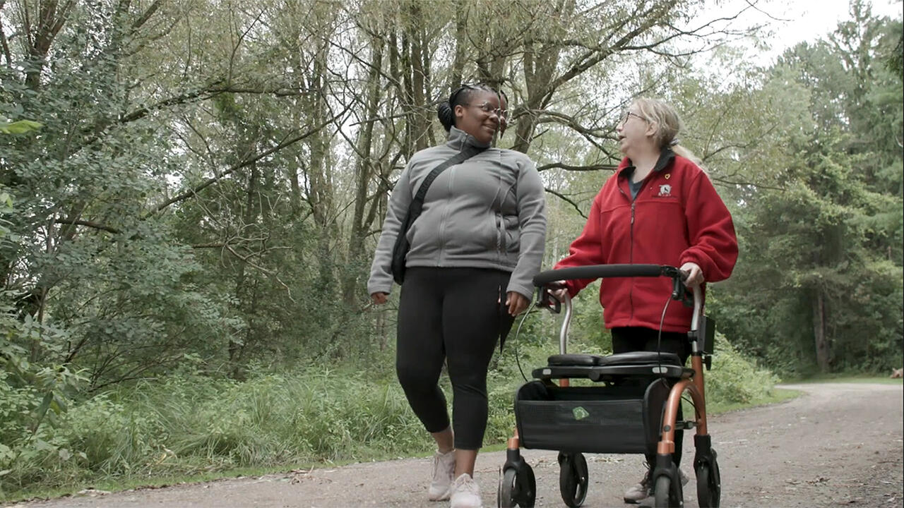 Two women walking on a nature trail, with one using a walker