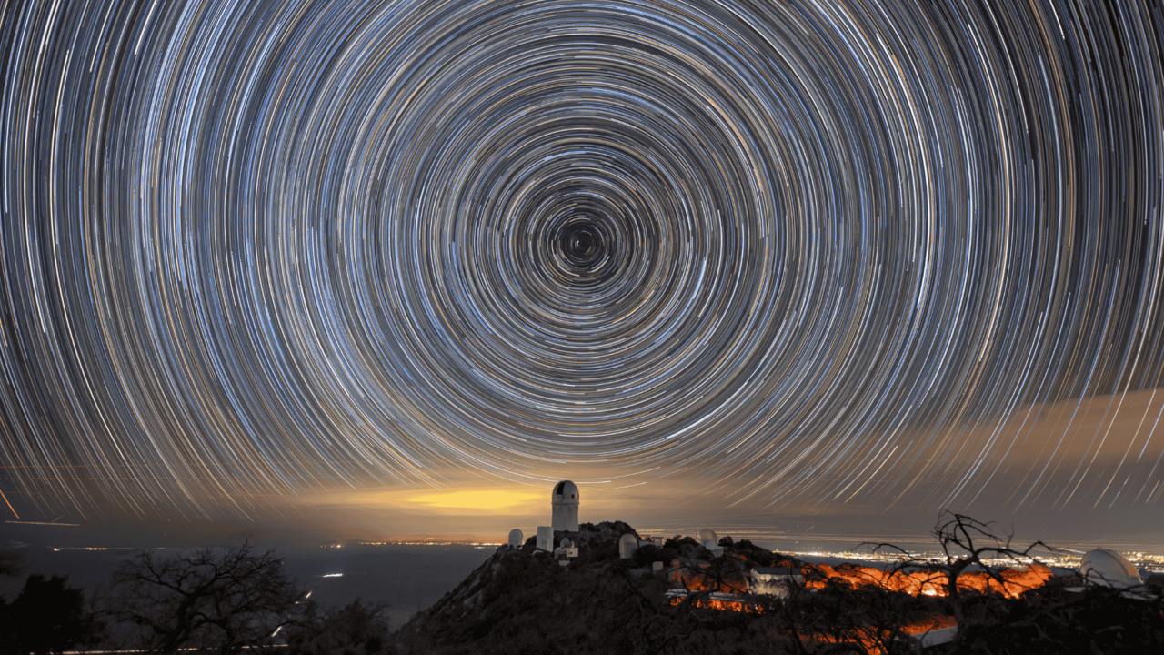 Circles of light on the night sky. A telescope dome atop a mountain is below the center of the circle.