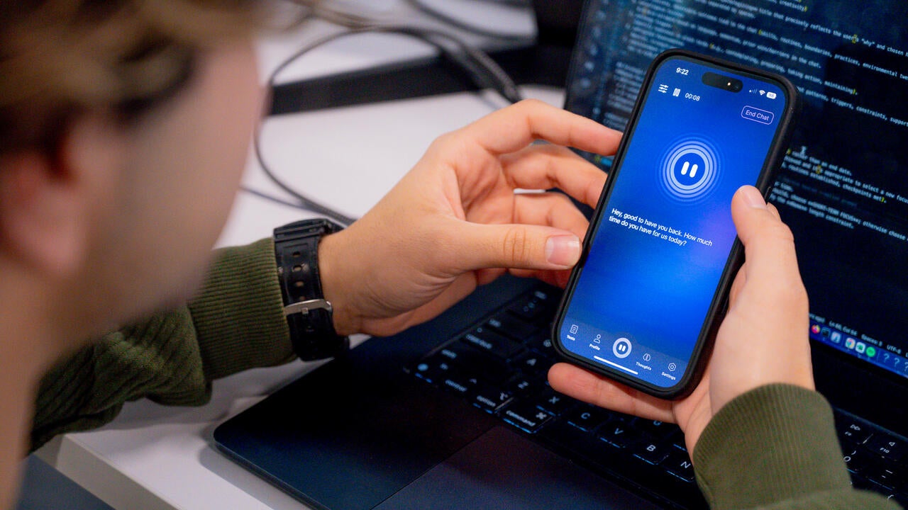 Rastin Rassoli holding a mobile phone with screen showing Doro AI interface and a laptop with coding in the background