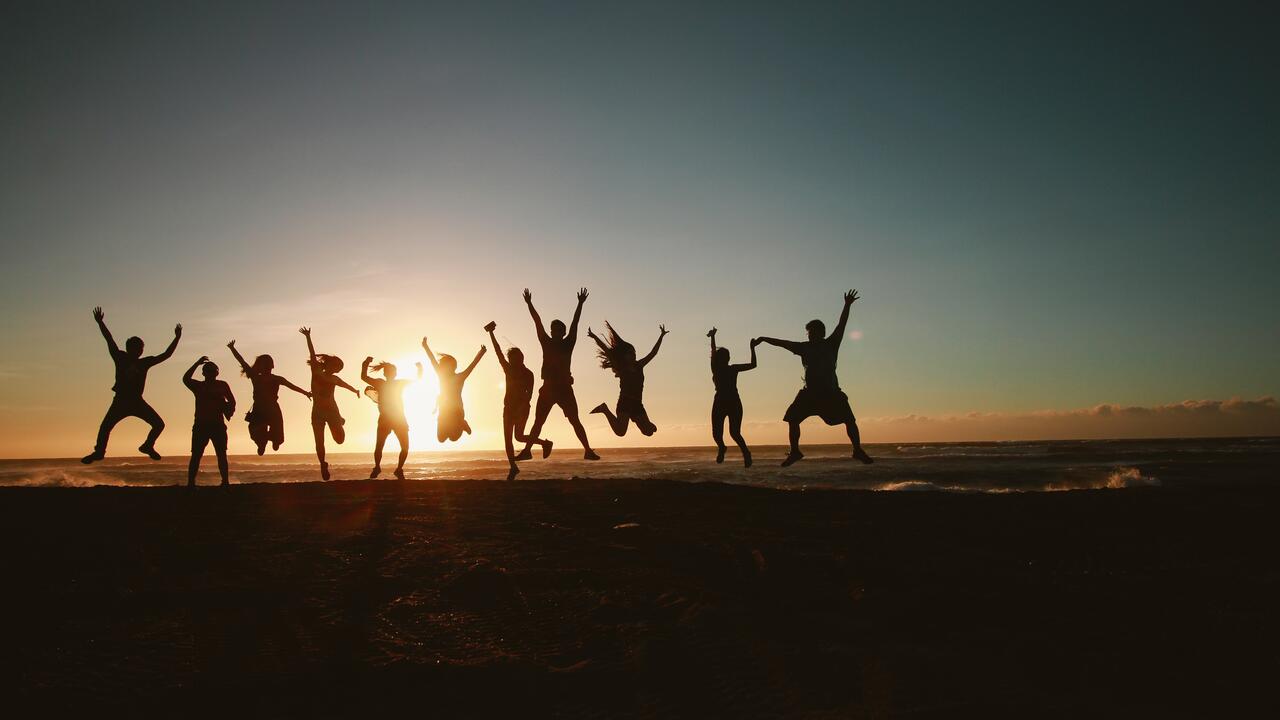 Shadowed group of friends holding hands in mid-air jump with sunset background